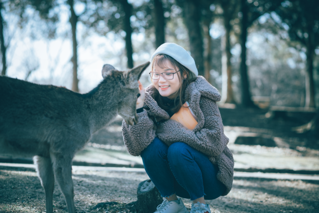 Smiling woman in warm clothing lovingly interacting with a deer in an outdoor park.