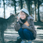Smiling woman in warm clothing lovingly interacting with a deer in an outdoor park.