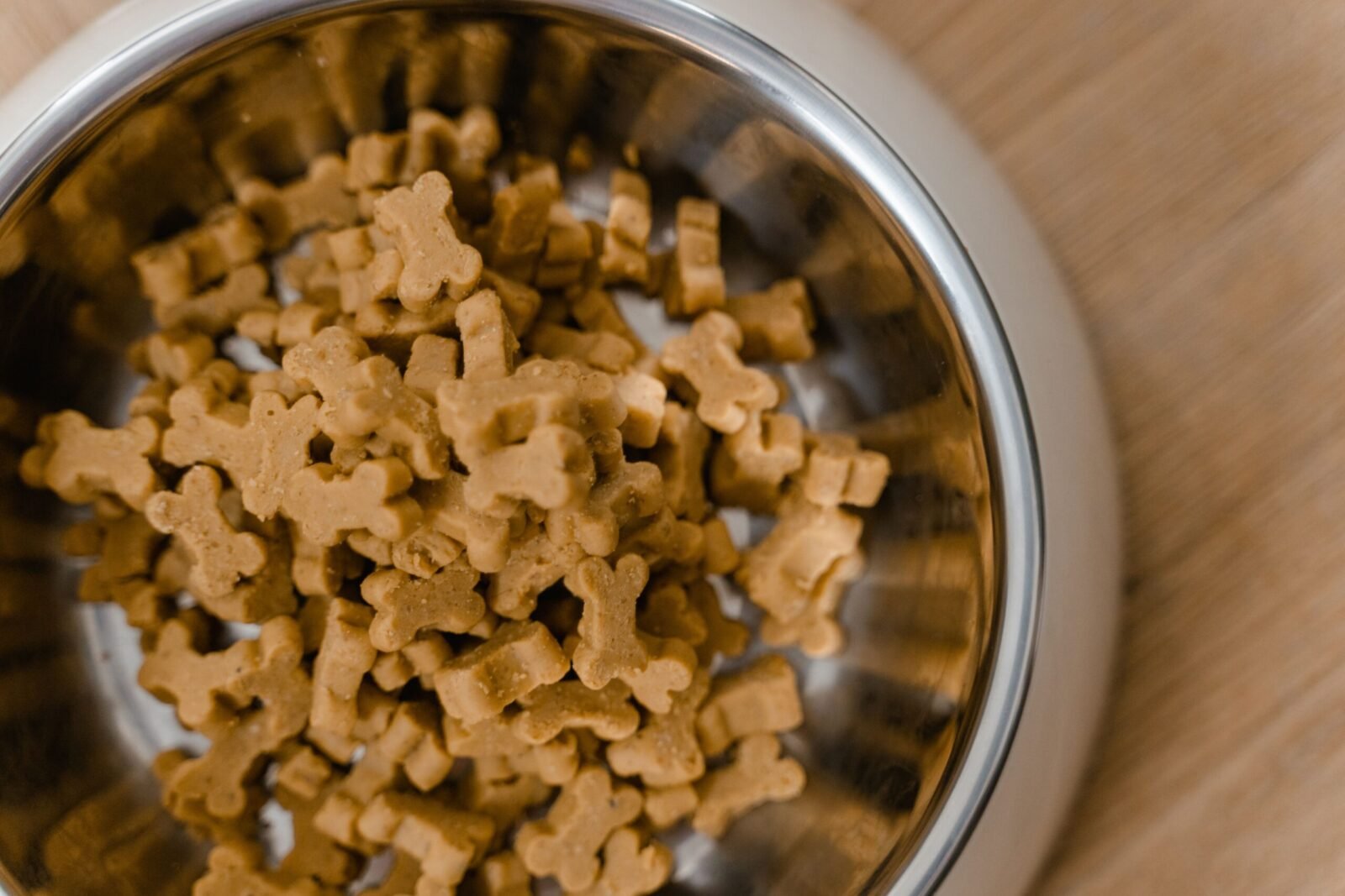 A close-up view of dog bone-shaped biscuits in a stainless steel bowl on a wooden surface.